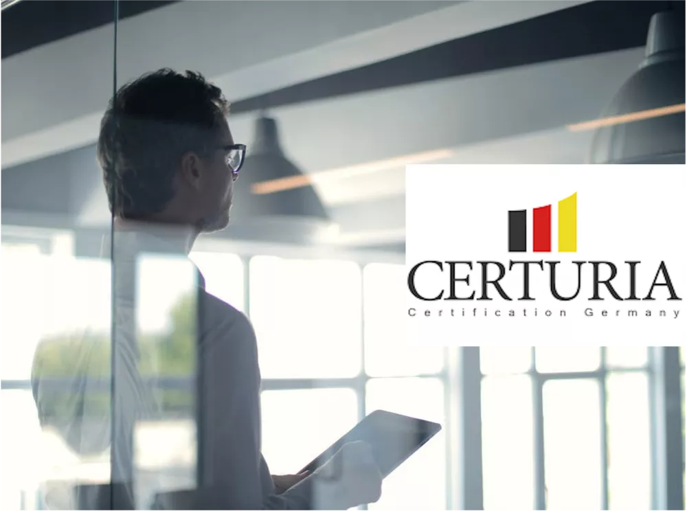 Ein Geschäftsmann in einem modernen Bürogebäude schaut aus einem Fenster, während er ein Tablet hält. Rechts oben ist das Logo von Certuria Certification Germany zu sehen, das aus drei vertikalen Balken in Schwarz, Rot und Gelb besteht, gefolgt von dem Schriftzug 