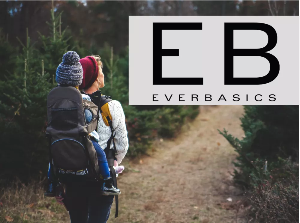 Das Bild zeigt einen Menschen mit einer Kinderkraxe und einem Kind darin bei einer Wanderung im Wald. Rechts oben ist das EB Everbasics Logo zu sehen.